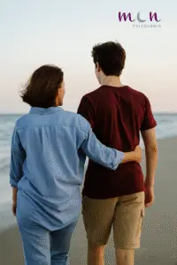 Madre caminando junto a su hijo adolescente por la orilla del mar, con el brazo sobre sus hombros, simbolizando el acompañamiento respetuoso, la confianza y el vínculo emocional positivo.
